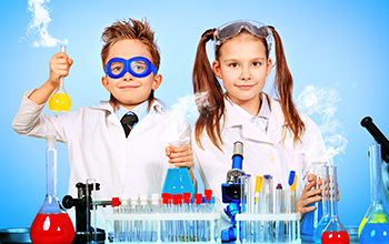 A boy wearing blue safety goggles and a girl wearing lab coats working with beakers in a lab