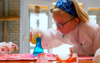 A young girl in a lab coat pouring yellow liquid into a blue Erlenmeyer flask.