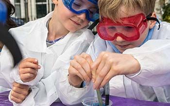 A girl and a boy wearing safety goggles and a mad science lab coat pouring liquid in a cup.