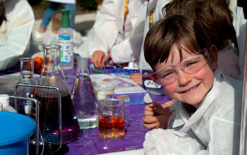 A smiling boy at a science booth, with beakers and other kids in the background.